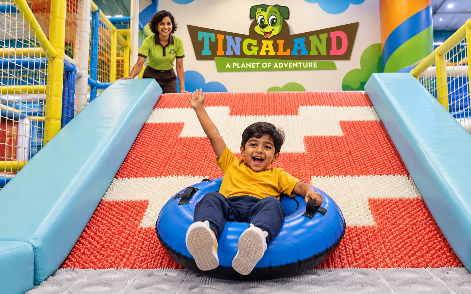 Boy riding a blue donut ring down the tingaland donut slide while a uniformed play attendant supervises