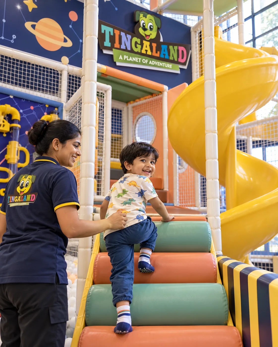 Toddler spiral slide 2 Toddler climbing padded steps toward the yellow spiral slide with an attendant